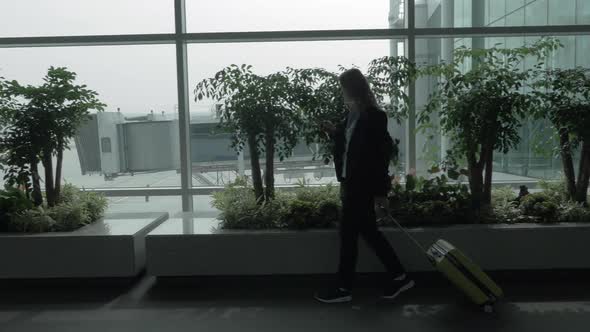 Girl with Cell and Suitcase Walking By the Window of Airport Terminal