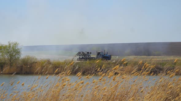 Tractor Drives Along the Shore of the Lake Against the Background of Smoke and Fields