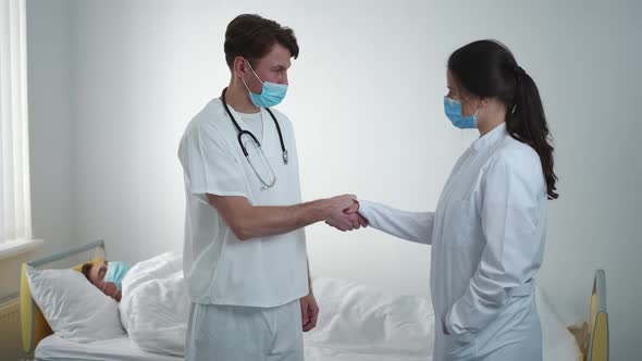 Intelligent Caucasian Doctors Shaking Hands Standing in Hospital Ward with Ill Patient