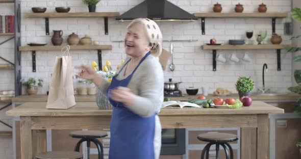 Portrait of Cheerful Female Retiree Dancing and Spinning with Cooking Utensils Indoors. Smiling