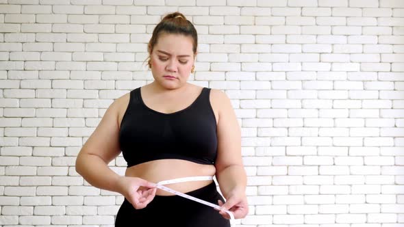Unhappy overweight woman in sportswear measuring waist with measure tape