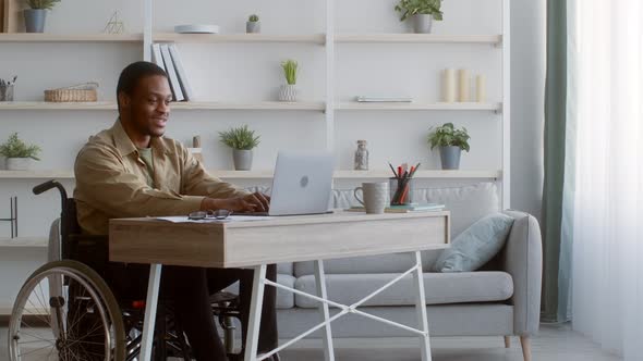 Disabled African Guy Working On Laptop Sitting In Wheelchair Indoors