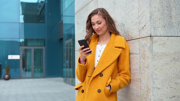 Business Woman Dressed Yellow Coat Standing Outdoors Corporative Building Background with Smartphone