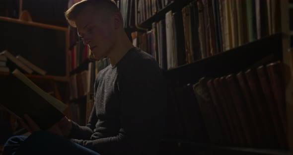 A Young Guy Is Sitting on the Floor Between the Bookshelves and Leafing Through a Book. Taken in