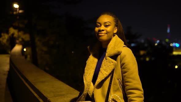 A Young Black Woman Smiles at the Camera As She Sits on a Low Wall in a Park in Urban Area at Night