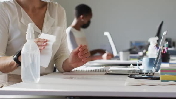 Two diverse female colleagues wearing face mask, sanitizing desk in office