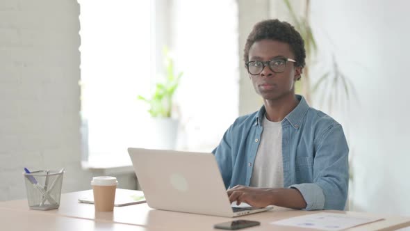 Young African Man Smiling at Camera While Using Laptop in Office