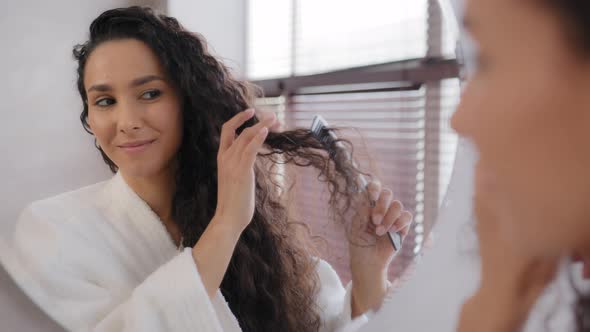 Closeup Happy Smiling Young Woman in Bathrobe Looking in Mirror Brushing Combing Long Curly Hair