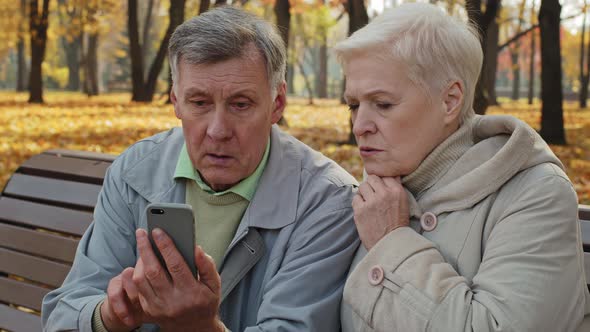 Upset Elderly Married Couple Sit on Bench in Autumn Park Read Bad News on Smartphone Worried Old