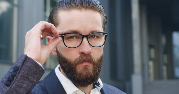 Bearded Businessman with Glasses Looking at Camera Near Modern Office Building