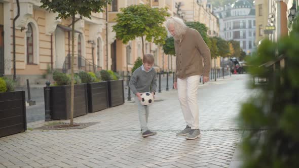 Happy Little Boy Running with Soccer Ball To Smiling Senior Man. Joyful Caucasian Grandson and