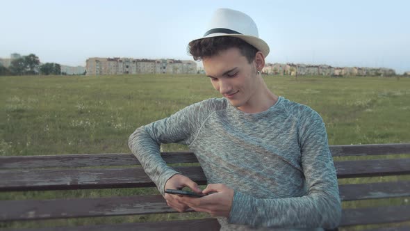 Guy in a Hat Who Sits on an Old City Bench with a Phone Reads Messages and Smiles Camera Tracking