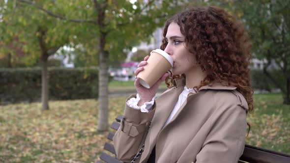 Autumn Young Beautiful Woman Drinks Coffee or a Hot Drink in the Park on a Bench