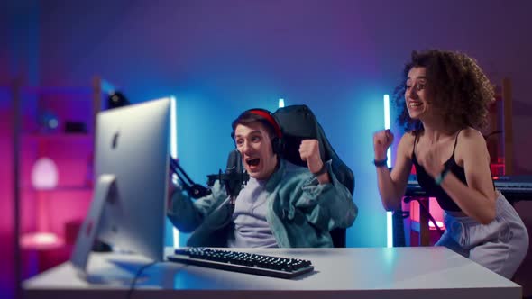 Excited Gamer Guy and Young Woman Wearing Headphones Playing Computer Game Neon Fashion Room Winner