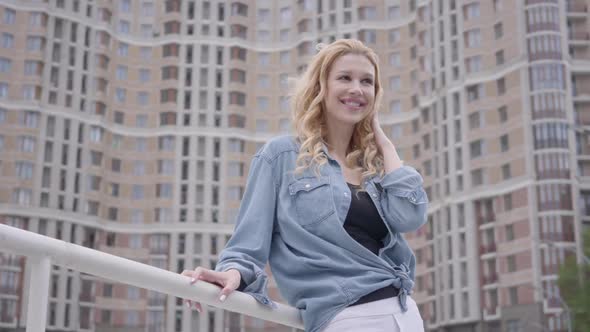 Pretty Confident Blond Woman in Jeans Jacket Posing in Front of Skyscraper, Looking Away Smiling