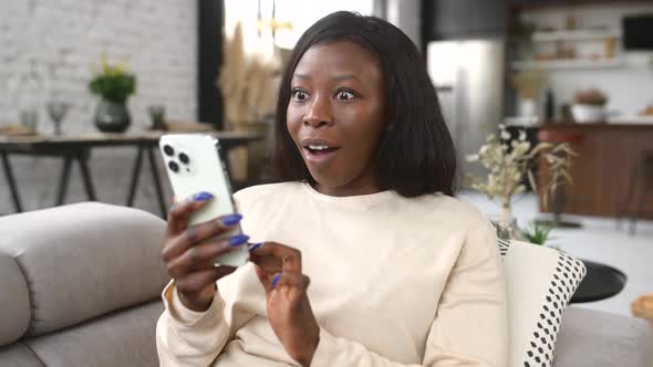 Excited Young Woman in Casual Wear Rejoices Sitting on the Couch and Holding Smartphone