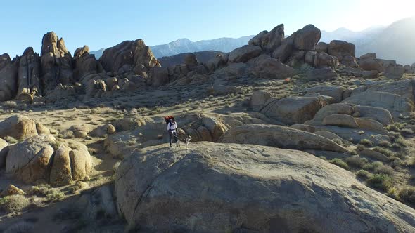 Aerial shot of a young man backpacker standing on a boulder with his dog in a desert mountain range