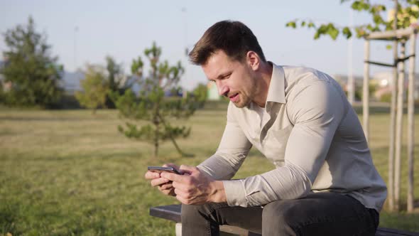 A Young Caucasian Man Plays a Game on a Smartphone As He Sits in a Park in an Urban Area