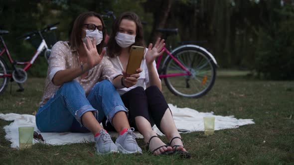 Two Young Women in Face Masks Sitting at the Park and Taking a Video Call