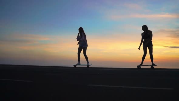 Two Girlfriend Girls in Shorts and Sneakers Ride Skateboards on the Slope Against the Beautiful Sky