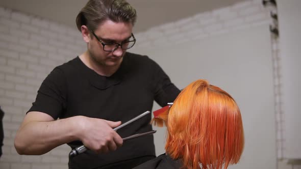 Male Professional Hair Dresser is Straightening Bright Red Woman's Hair Using a Hairstraightener in