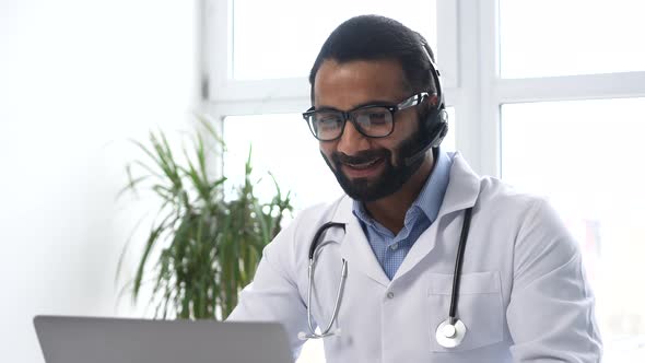 Indian Male Doctor with a Stethoscope in Gown Sitting at the Desk and Looking at the Laptop