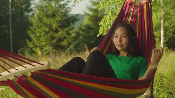 Carefree Lovely Asian Female Tourist in Hammock Enjoying Music in Headphones in Mountain Nature