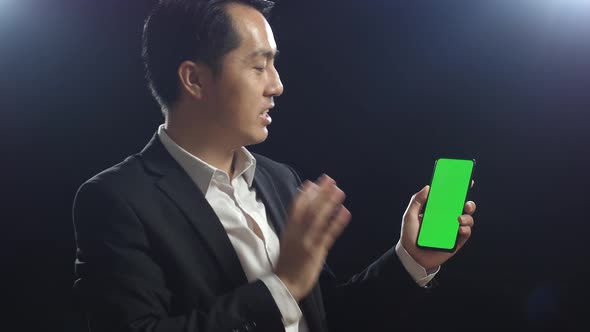 Asian Speaker Man In Business Suit Holding And Pointing Green Screen Smartphone While Speaking