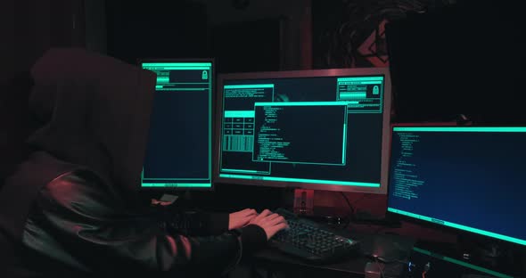 Hacker in a Hood Sits in Front of Computer Screens and Hacks Databases