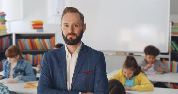 Young Male Teacher Standing in Classroom with Kids Sitting at Desk on Background