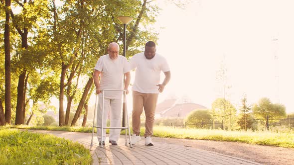 African-American caregiver is teaching disabled old man to walk with walker. Nurse and patient.