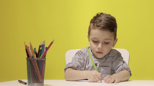 Smiling little boy at the table draw with crayons for mum on yellow background. Creativity concept