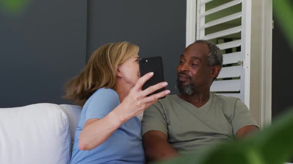 Mixed race senior couple taking a selfie on smartphone while sitting on the couch at home