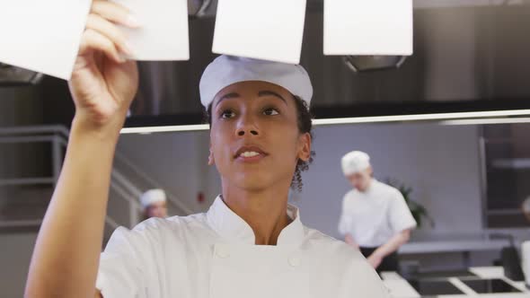 African American female chef working in a restaurant kitchen checking orders, with colleagues workin