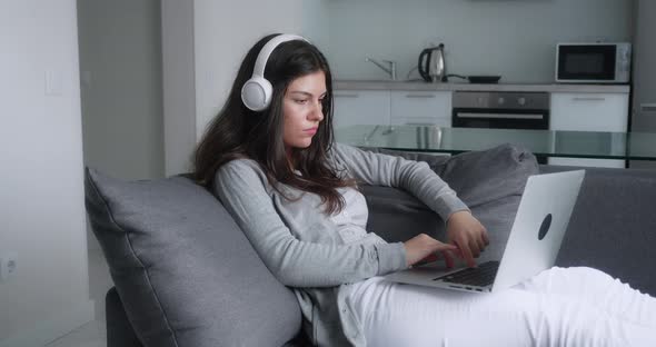 Focused Young Adult Woman in Headphones Sitting in Cozy Living Room on Couch Holding Using Laptop