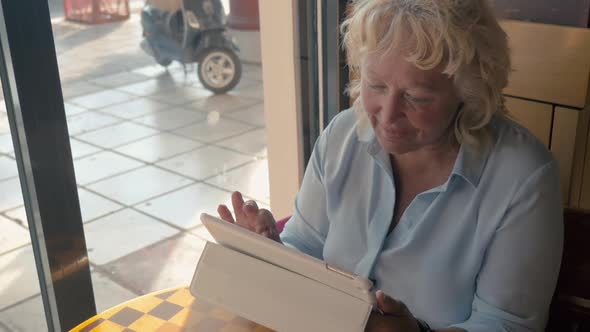 Mature Woman Sitting in a Cafe and Enjoys Her Tablet., Outside the Window the Summer, Sunny Day