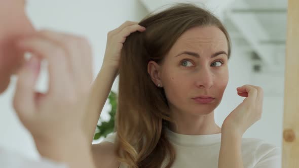 Sad Upset Woman Standing By Mirror Looking at Matted Fragile Strand in Her Hand Thinking About