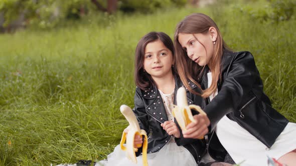 Cheerful Little Sisters Sitting Closely to Each Other Holding Bananas in Hands