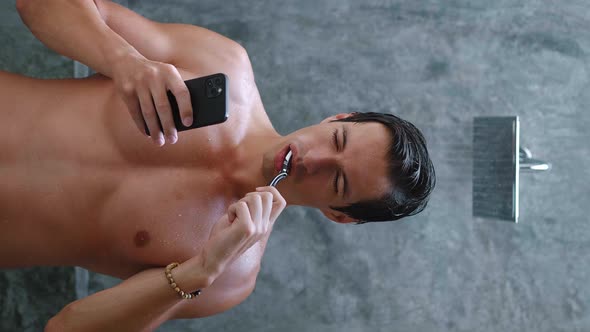 Vertical Video Portrait of a Man Brushing His Teeth and Using a Smartphone While Standing in the