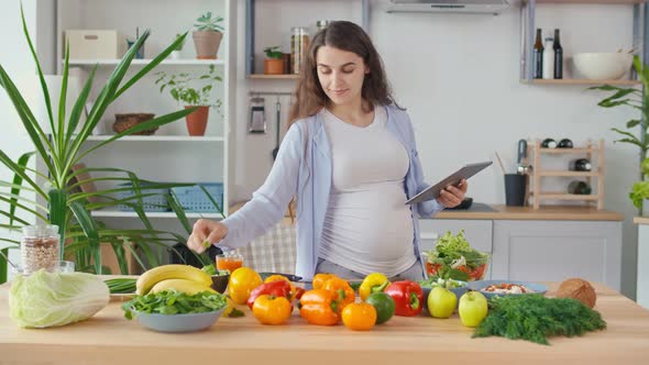 A Pregnant Woman Looks for a Recipe on a Tablet And Makes a Salad in a Bright