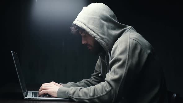 Side View of Young Bearded Programmer Hacking Online Site or Password at Desk in Dark Room