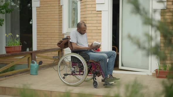 Absorbed Handicap African American Man Reading Book Sitting in Wheelchair on Porch Outdoors