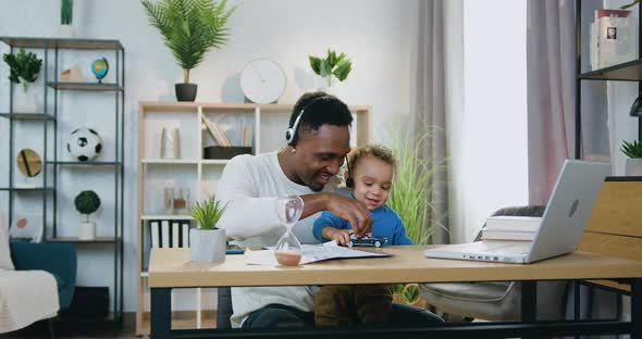 Black-Skinned Dad in Headset Sitting at His Workplace Together with Small Son and Playing
