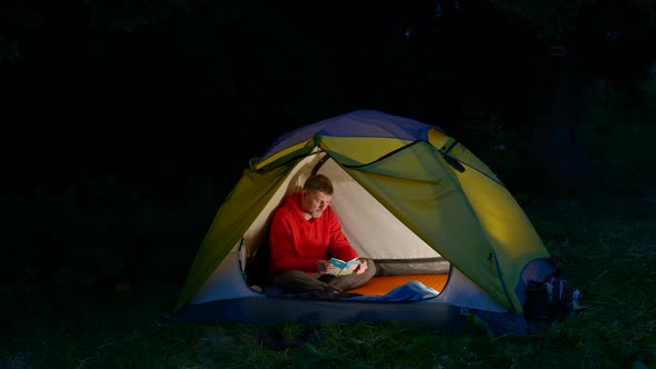 A Man Leafs Through a Notebook in a Tent in the Night Forest