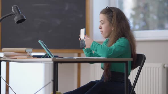 Side View Smiling Happy Little Person Sitting at Desk Showing Smartphone at Tablet Video Chat