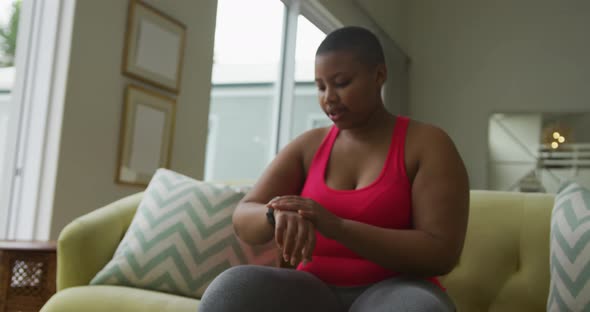 Happy african american plus size woman practicing yoga, using smartwatch in living room