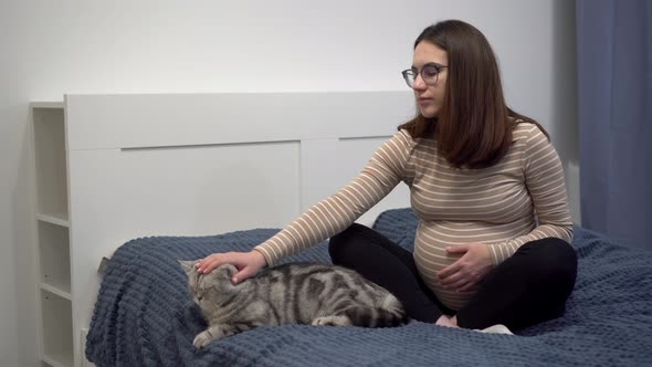 A Young Pregnant Woman with Glasses is Stroking a Cat