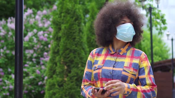 Portrait Afro American Woman with an Afro Hairstyle in a Protective Medical Mask with a Smartphone