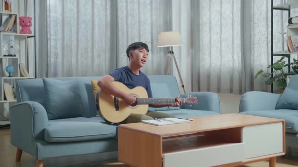 Asian Boy Composer With Notebook On Table Playing Guitar At Home