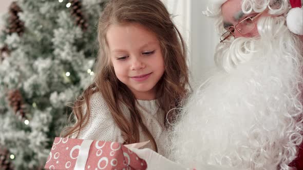 Santa Claus Tying a Ribbon Bow on a Gift for a Little Girl Sitting Near the Christmas Tree at Home